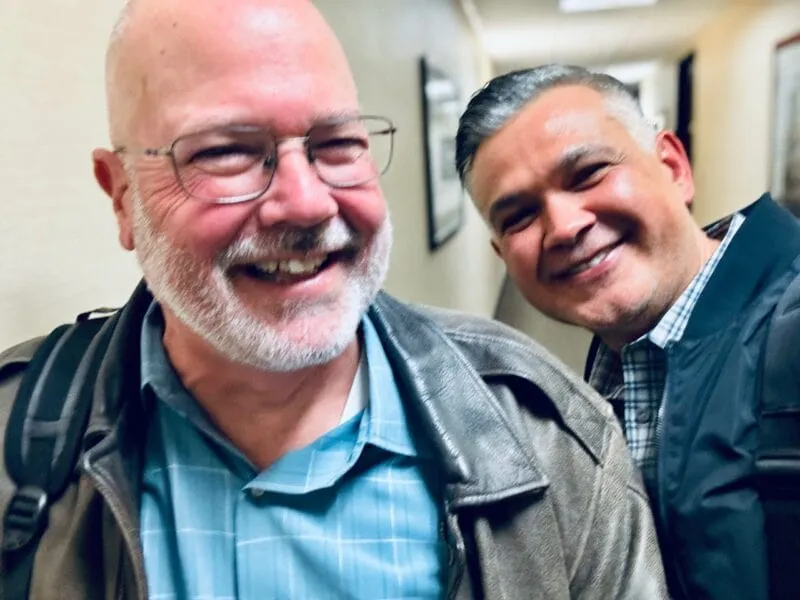 Two cheerful men smiling in a hallway, one wearing glasses and a leather jacket, the other in a checkered shirt and jacket, posing closely for a selfie.