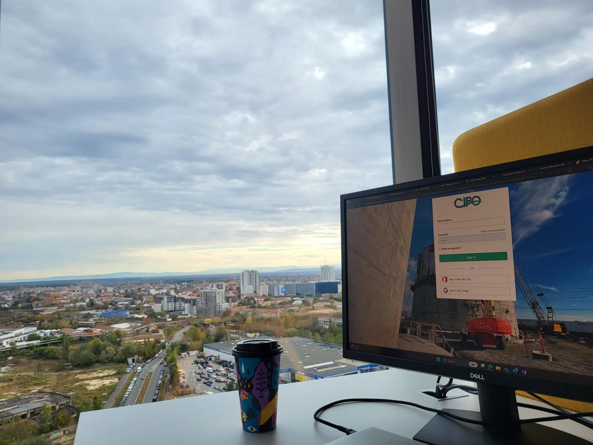 Office desk with a colorful coffee cup and a computer monitor displaying the CIPO login page, with a scenic city view and cloudy sky visible through a large window.