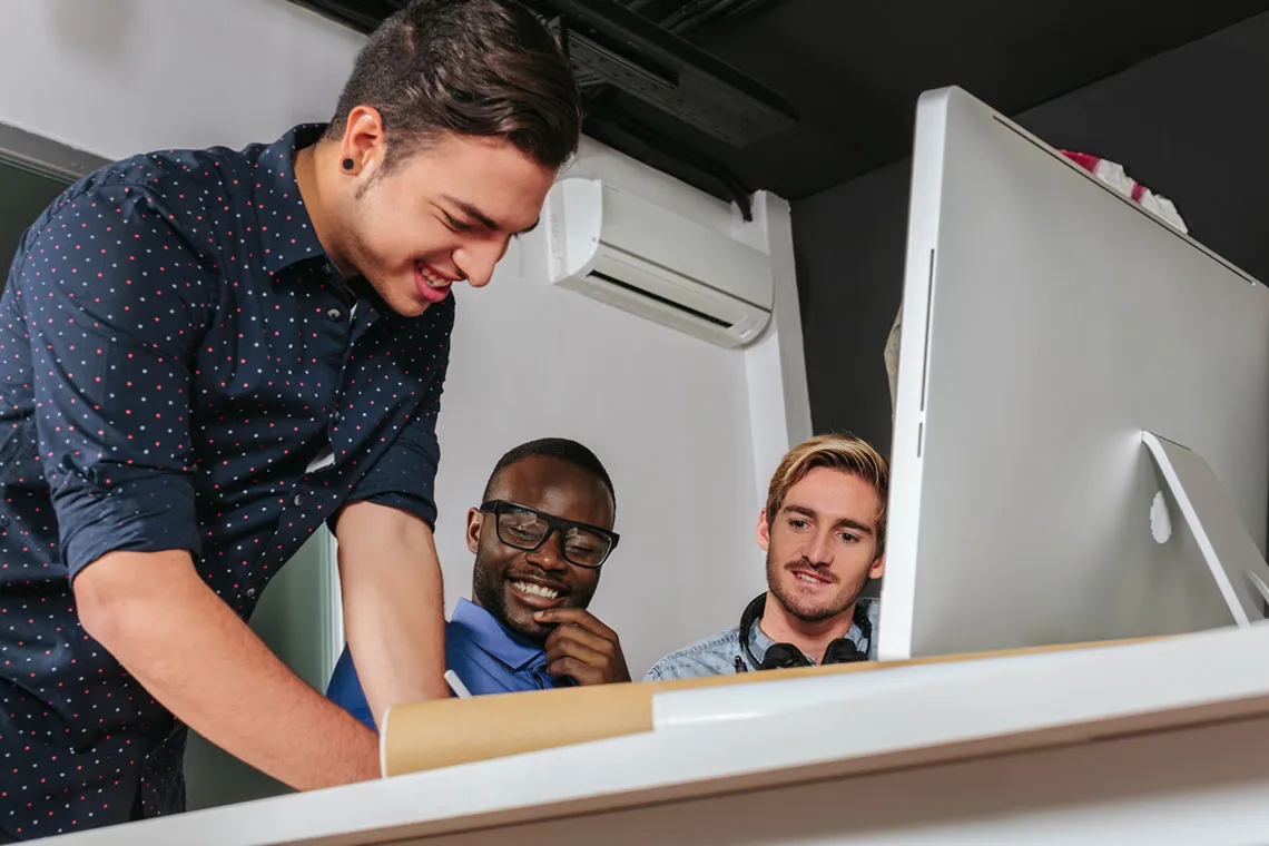 Three young professionals collaborating at a desk, with one standing and pointing at a large sheet while the others observe a computer monitor.