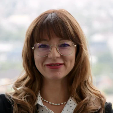 Smiling woman with light brown wavy hair, glasses, and a pearl necklace, wearing a polka-dotted blouse and dark blazer, standing in front of a blurred cityscape background.