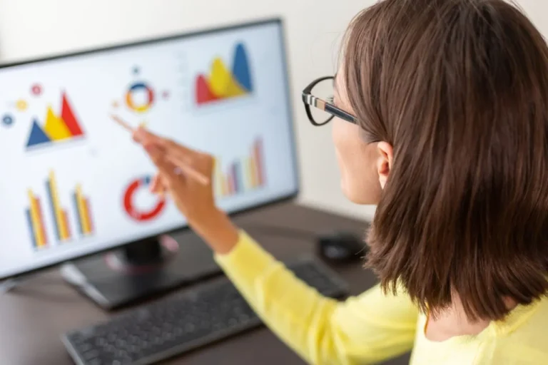 Woman in glasses analyzing colorful graphs and charts on a computer screen with a pencil.
