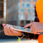 Close-up of a construction worker using a tablet on-site, with a blurred building in the background.