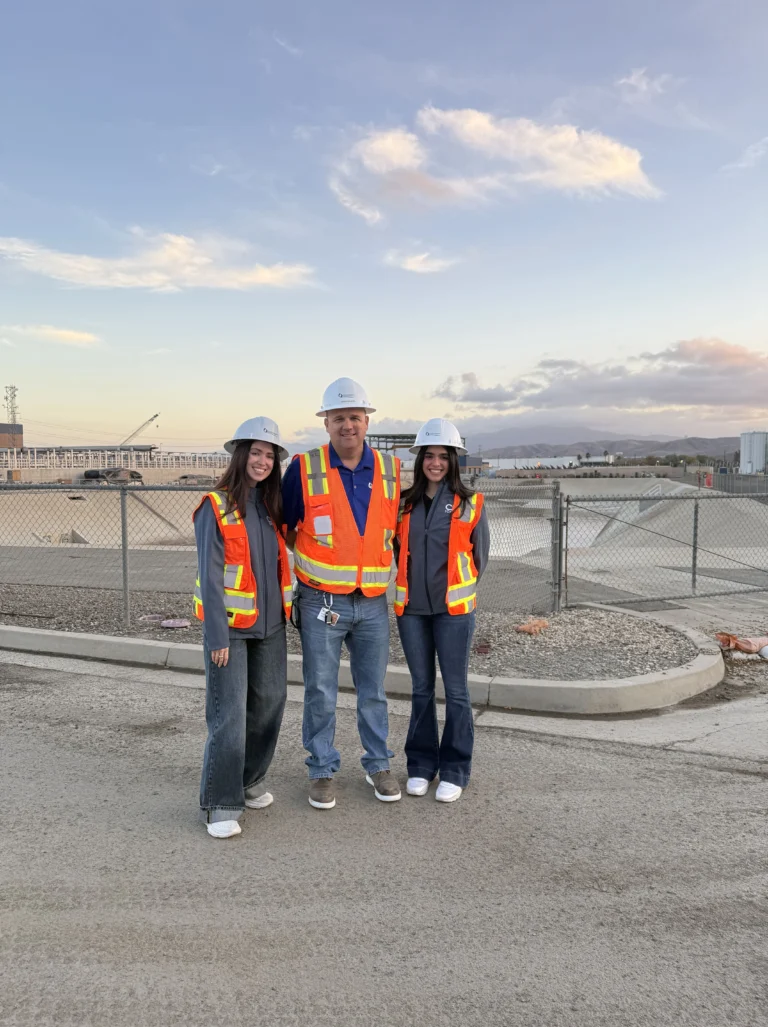 Three people wearing hard hats and safety vests standing at a construction site with a sunset sky in the background.