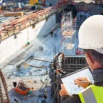 Construction engineer with hard hat and safety vest reviewing architectural plans on a tablet at an active building site.