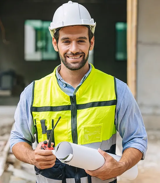 Smiling construction engineer wearing a white hard hat and neon safety vest, holding blueprints and a walkie-talkie at an active building site