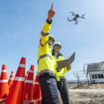 Two engineers in high visibility jackets and helmets operate a drone using a laptop amidst a stack of traffic cones on a construction site. The scene highlights advanced technology and teamwork.