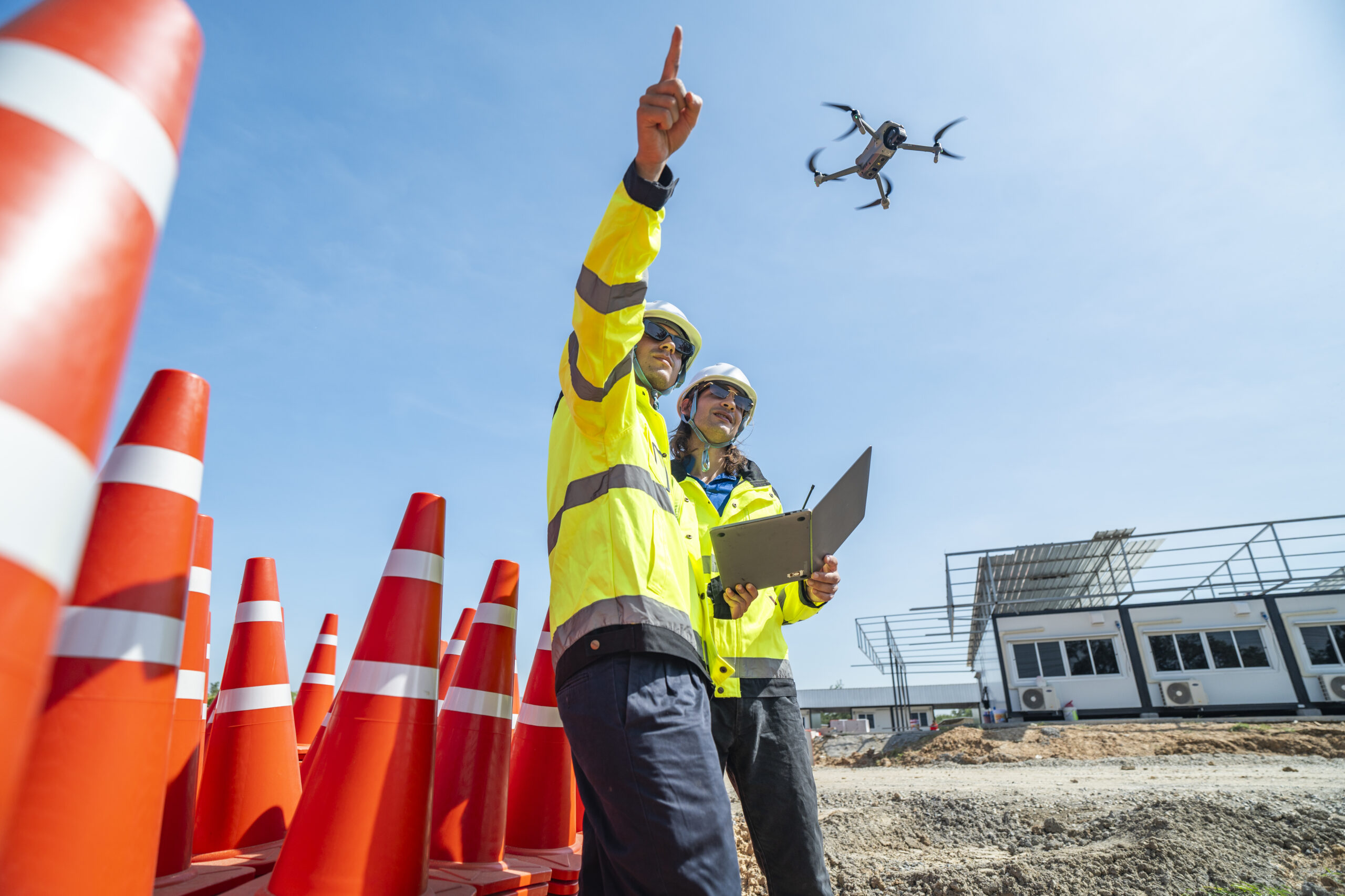 Two engineers in high visibility jackets and helmets operate a drone using a laptop amidst a stack of traffic cones on a construction site. The scene highlights advanced technology and teamwork.