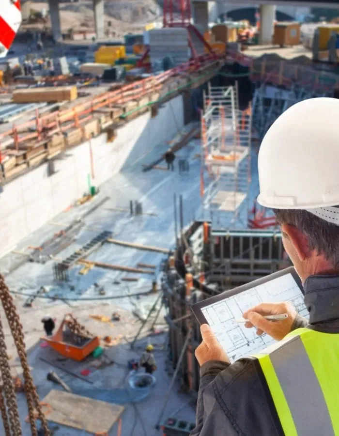 Construction engineer with hard hat and safety vest reviewing architectural plans on a tablet at an active building site.
