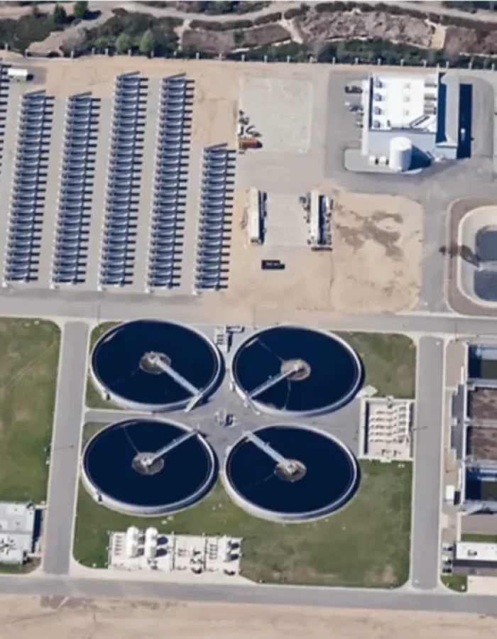 Aerial view of a large wastewater treatment facility. The image shows multiple circular clarifiers, rectangular treatment basins, a solar panel array, and administrative or industrial buildings. There is also a large settling pond and various service roads and green areas across the complex.